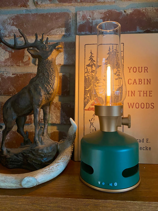 A green KooKoo MoriMori Light & Sound Lantern with a nostalgic design and glass chimney glows on a wooden surface next to a deer figurine and a book.