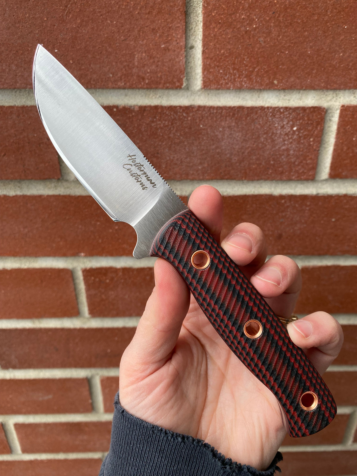 Hesterman Outback Hunter Knife with satin-finished blade, etched branding, red-black handle, and jimping, held against a brick wall.