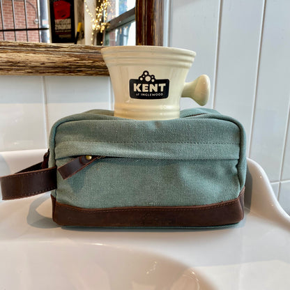 Dopp Kit & Shaving Mug Bundle featuring a light green canvas bag with leather accents and a cream Kent of Inglewood shaving mug on a bathroom sink.