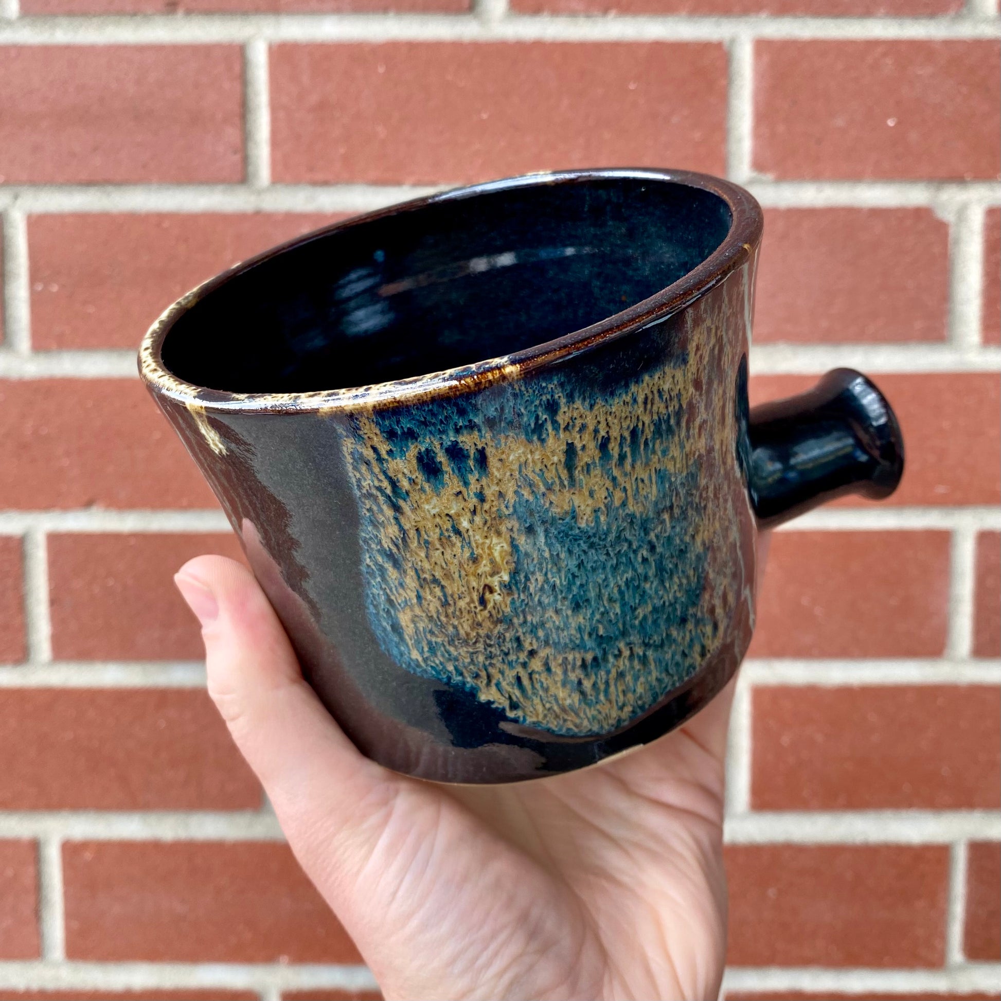 Sosan Fong - Apothecary Mug held against a red brick wall, showcasing its hand-thrown ceramic design with a unique, drippy glaze and short handle.