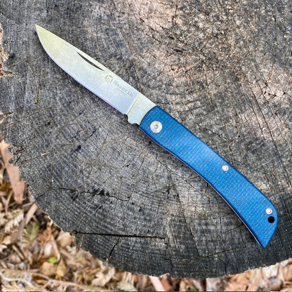 Maserin Scout 163 folding pocket-knife with a stone-washed steel blade and textured blue handle, resting on a tree stump, showcasing quality craftsmanship and durability.