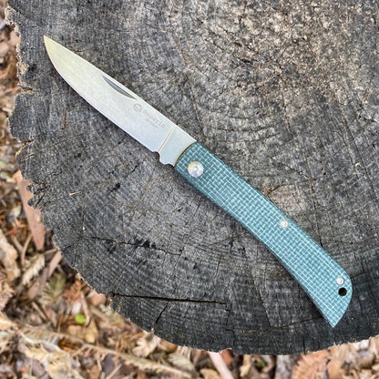 Maserin Scout 163 folding pocket knife with green-textured handle sits open on a tree stump, showcasing its sleek design and high-quality D2 steel blade.