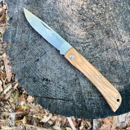 Maserin Scout 163 folding knife on a tree stump, showcasing an open D2 steel blade and light olive wood handle, ideal for everyday carry and outdoor tasks.