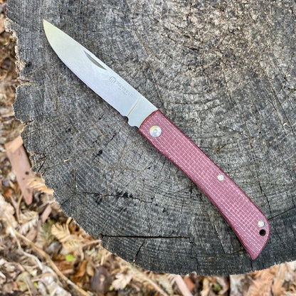 Maserin Scout 163 folding pocket knife with a textured red handle and open stainless-steel drop-point blade, displayed on a tree stump.
