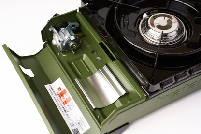 A close-up of an Iwatani Cassette Konro "Tough Maru" Portable Stove in green with its lid open, displaying the burner, gas cartridge compartment, and a label featuring usage instructions.