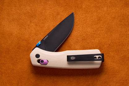 James Carter folding pocket knife with black drop-point blade, light G10 handle, blue thumb-stud, purple lock-slider, and black pocket clip, on orange suede.