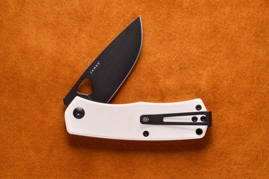James Folsom pocket knife with matte-black drop-point blade, white handle scales, and reversible deep-carry clip, open on a suede background.