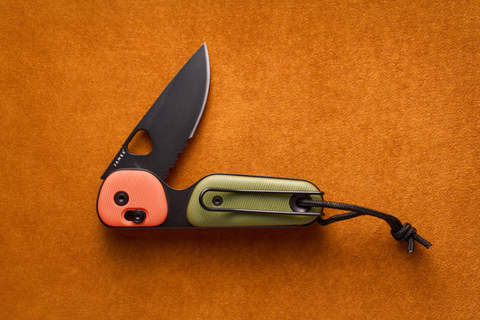 The James Redstone—a folding pocket knife featuring a partially serrated black blade, an orange and green handle, and a black lanyard, designed for everyday carry—is displayed on an orange textured surface.