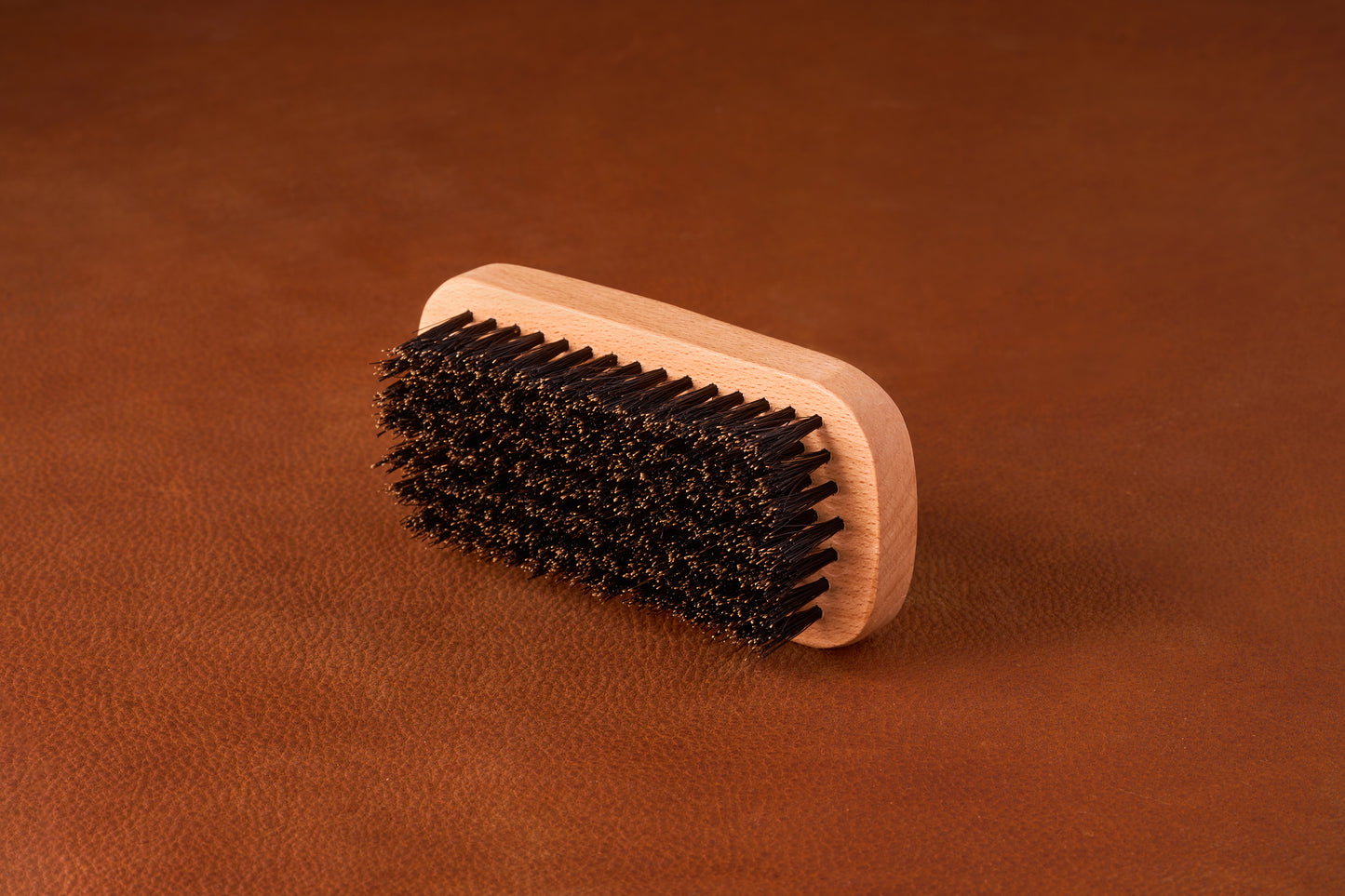 Kent Of Inglewood Boar Bristle Beard Brush on a leather surface, featuring a light wood handle and dense dark bristles for beard grooming and skin stimulation.