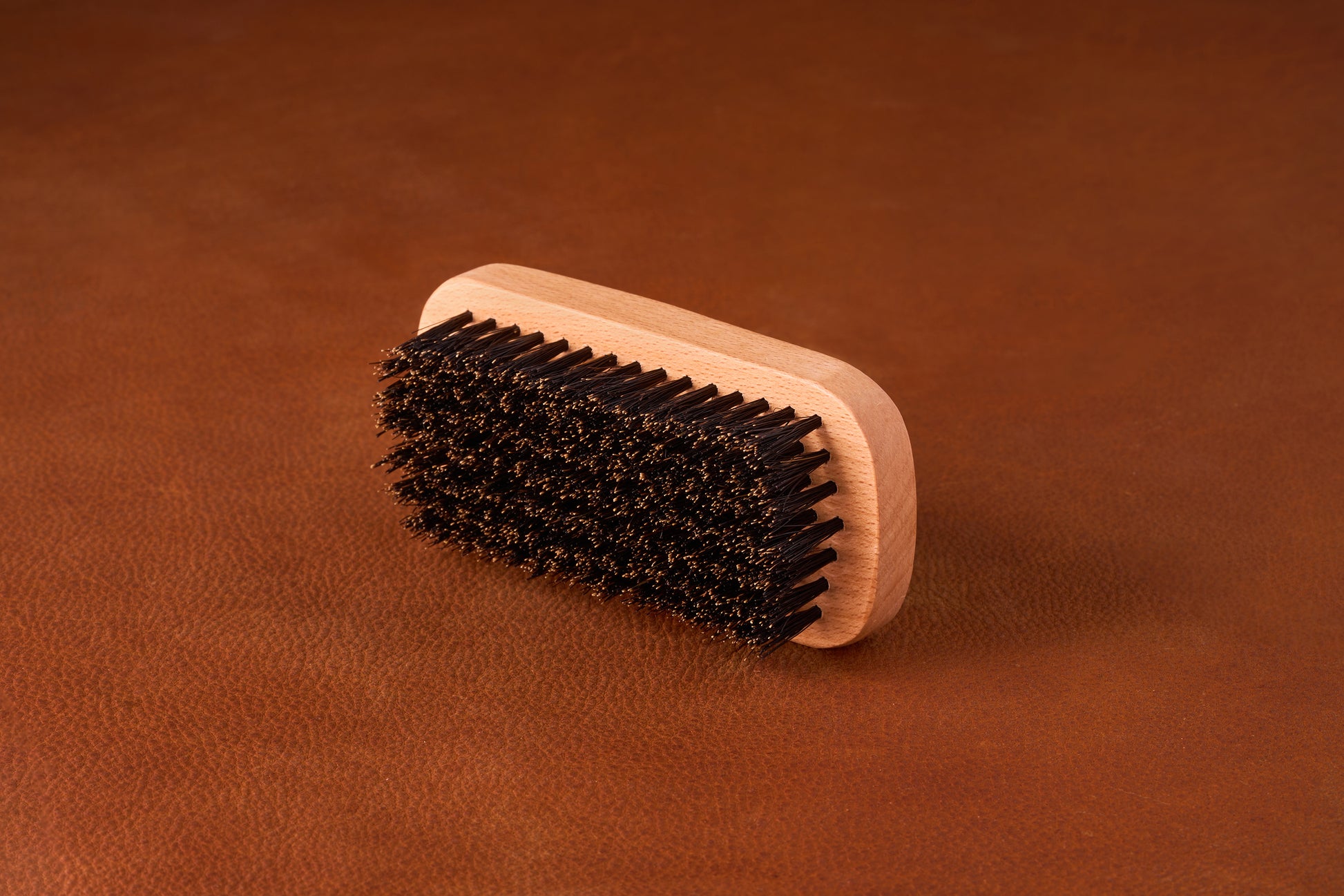 Kent Of Inglewood Boar Bristle Beard Brush on a leather surface, featuring a light wood handle and dense dark bristles for beard grooming and skin stimulation.