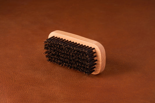 Kent Of Inglewood Boar Bristle Beard Brush on a leather surface, featuring a light wood handle and dense dark bristles for beard grooming and skin stimulation.