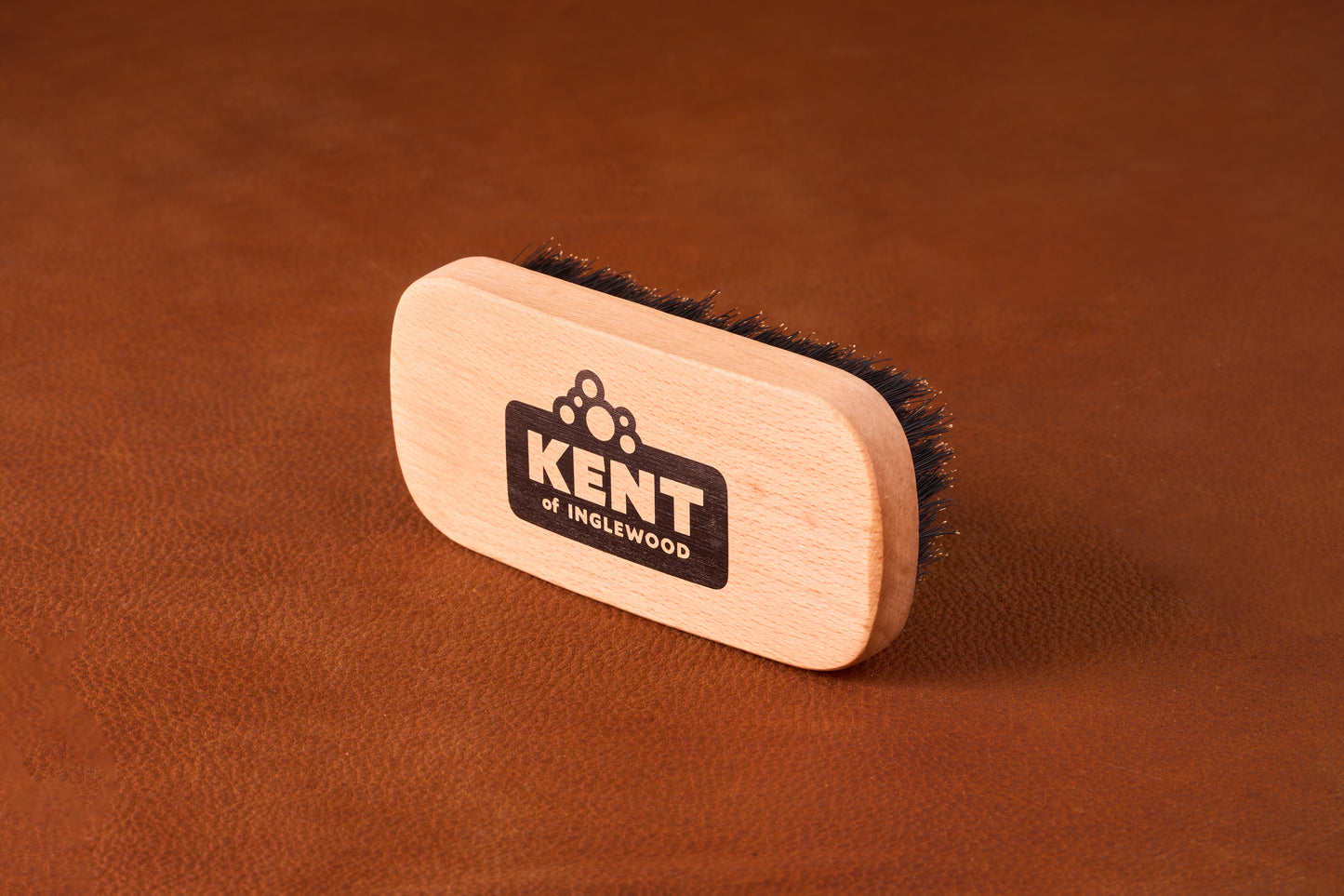 Kent Of Inglewood Boar Bristle Beard Brush on leather background, featuring dense dark bristles and a branded wooden handle for beard grooming and maintenance.