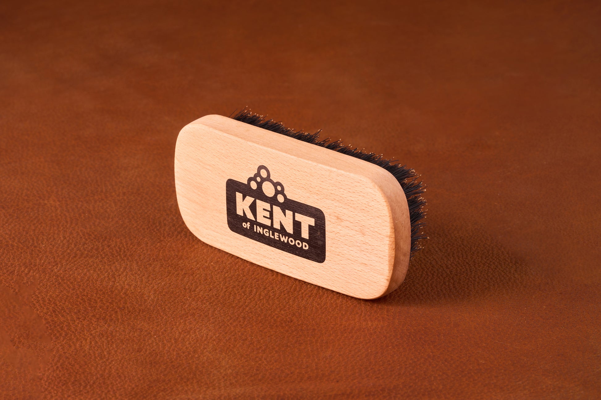 Kent Of Inglewood Boar Bristle Beard Brush on leather background, featuring dense dark bristles and a branded wooden handle for beard grooming and maintenance.