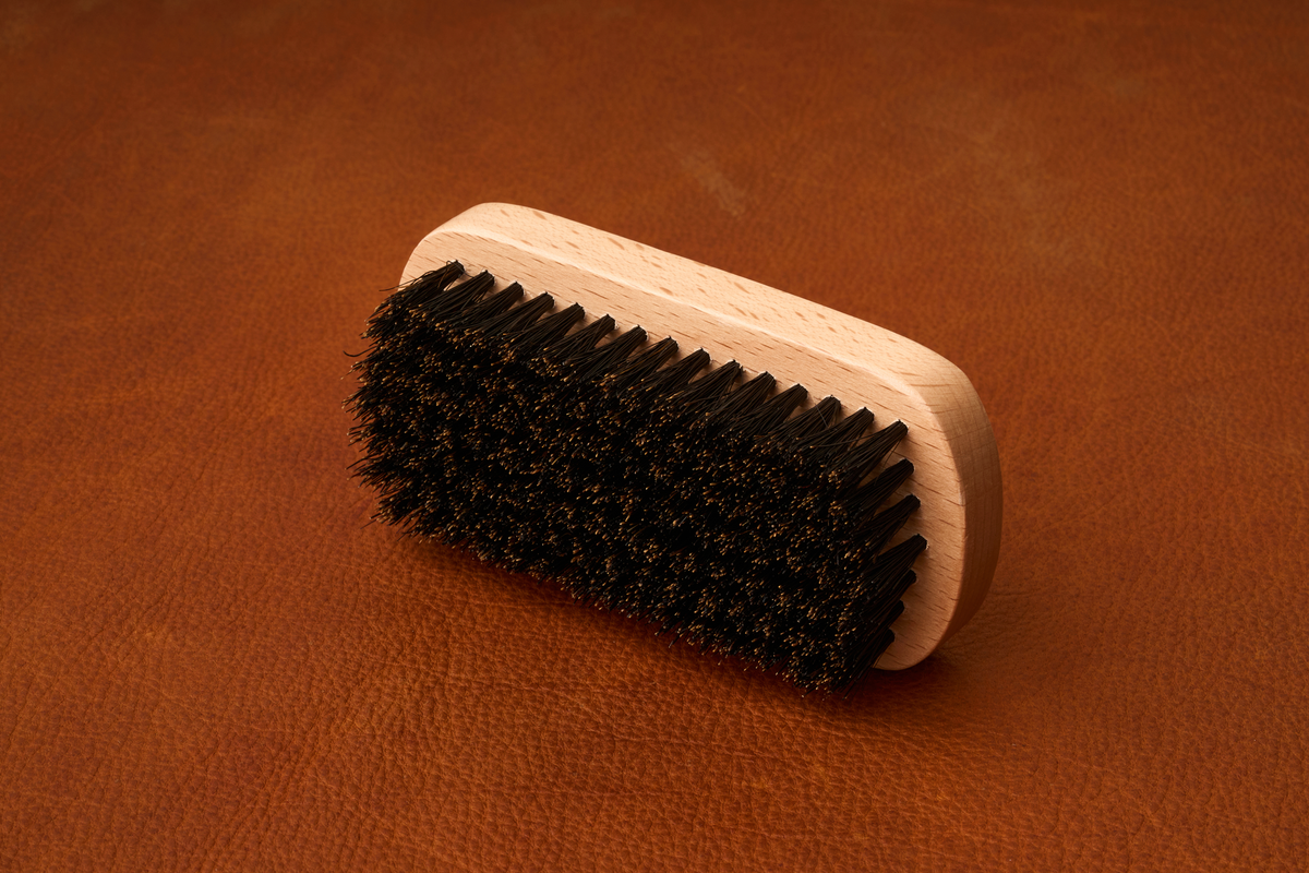 The Kent Of Inglewood Boar Bristle Beard Brush, featuring black bristles and a wooden handle, rests on a brown leather surface.