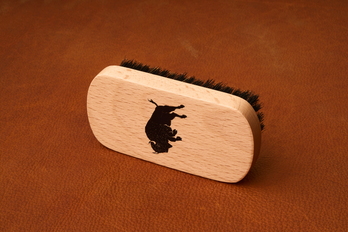 The Kent Of Inglewood Boar Bristle Beard Brush, featuring black boar bristles and a wooden handle printed with a black bull silhouette, rests on a brown leather surface—perfect for beard grooming.