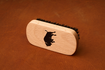 The Kent Of Inglewood Boar Bristle Beard Brush, featuring black boar bristles and a wooden handle printed with a black bull silhouette, rests on a brown leather surface—perfect for beard grooming.