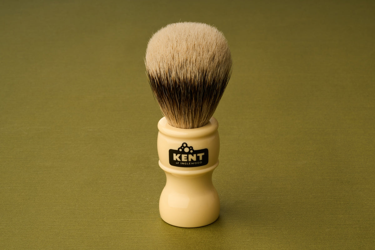 The Kent of Inglewood Silvertip Badger Hair Shaving Brush, with its cream-colored handle labeled “Kent” and dense, soft silvertip bristles, stands upright on a green textured surface.