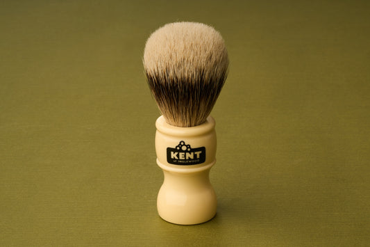 The Kent of Inglewood Silvertip Badger Hair Shaving Brush, with its cream-colored handle labeled “Kent” and dense, soft silvertip bristles, stands upright on a green textured surface.