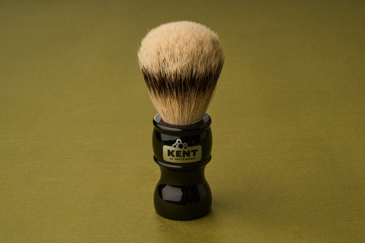 The Kent of Inglewood Silvertip Badger Hair Shaving Brush, featuring light bristles and a black handle labeled KENT, stands upright on an olive-green surface.