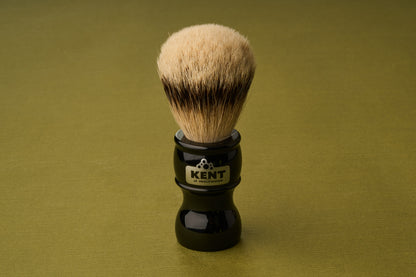 The Kent of Inglewood Silvertip Badger Hair Shaving Brush, featuring light bristles and a black handle labeled KENT, stands upright on an olive-green surface.
