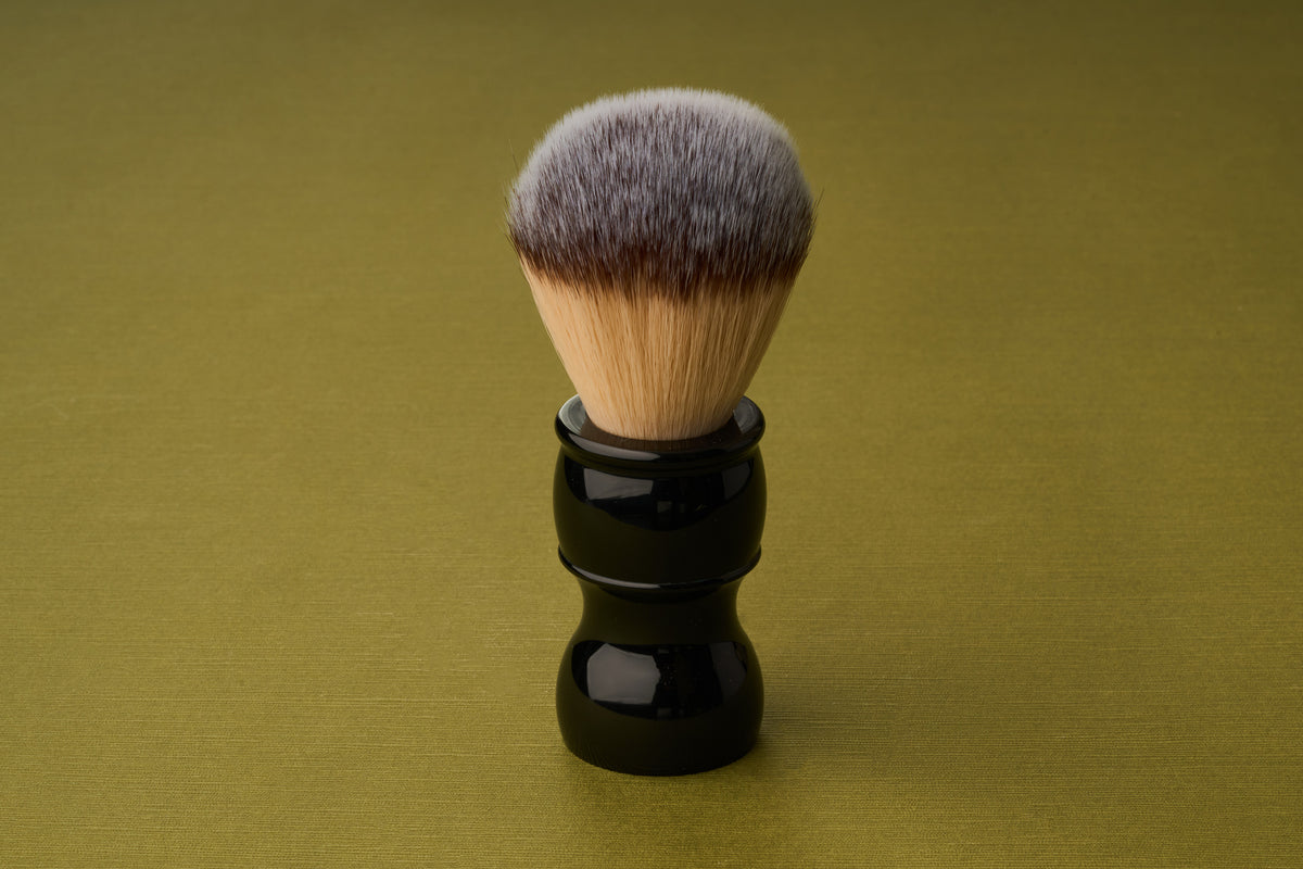 The Kent of Inglewood Synthetic Fibre Shaving Brush, featuring a black handle and dense light brown bristles, stands upright on a plain olive green background.
