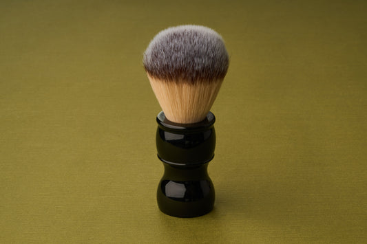 The Kent of Inglewood Synthetic Fibre Shaving Brush, featuring a black handle and dense light brown bristles, stands upright on a plain olive green background.
