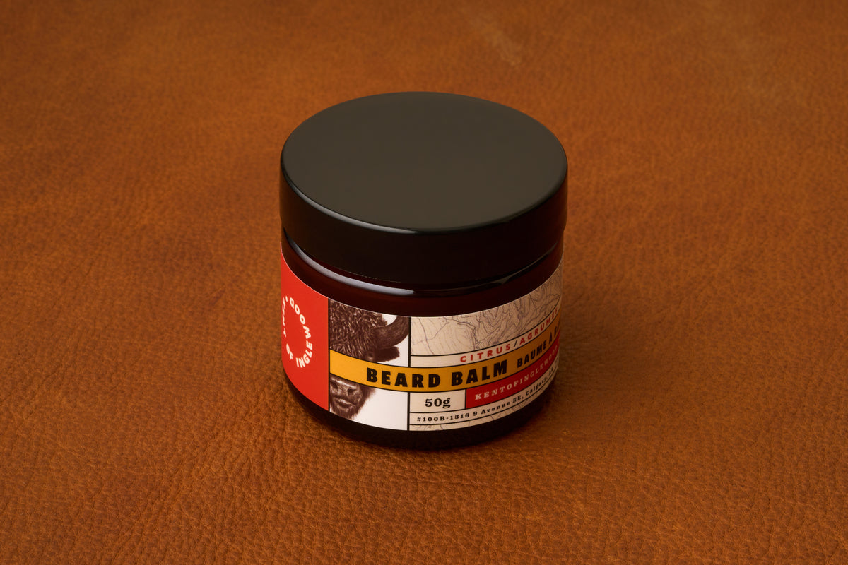 A jar of Kent of Inglewood Citrus Beard Balm with a brown label rests on a brown leather surface. The label displays an animal illustration and text noting that it contains 50g of beard balm, specially formulated to provide lasting beard moisture.