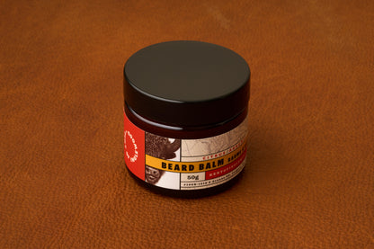 A jar of Kent of Inglewood Citrus Beard Balm with a brown label rests on a brown leather surface. The label displays an animal illustration and text noting that it contains 50g of beard balm, specially formulated to provide lasting beard moisture.