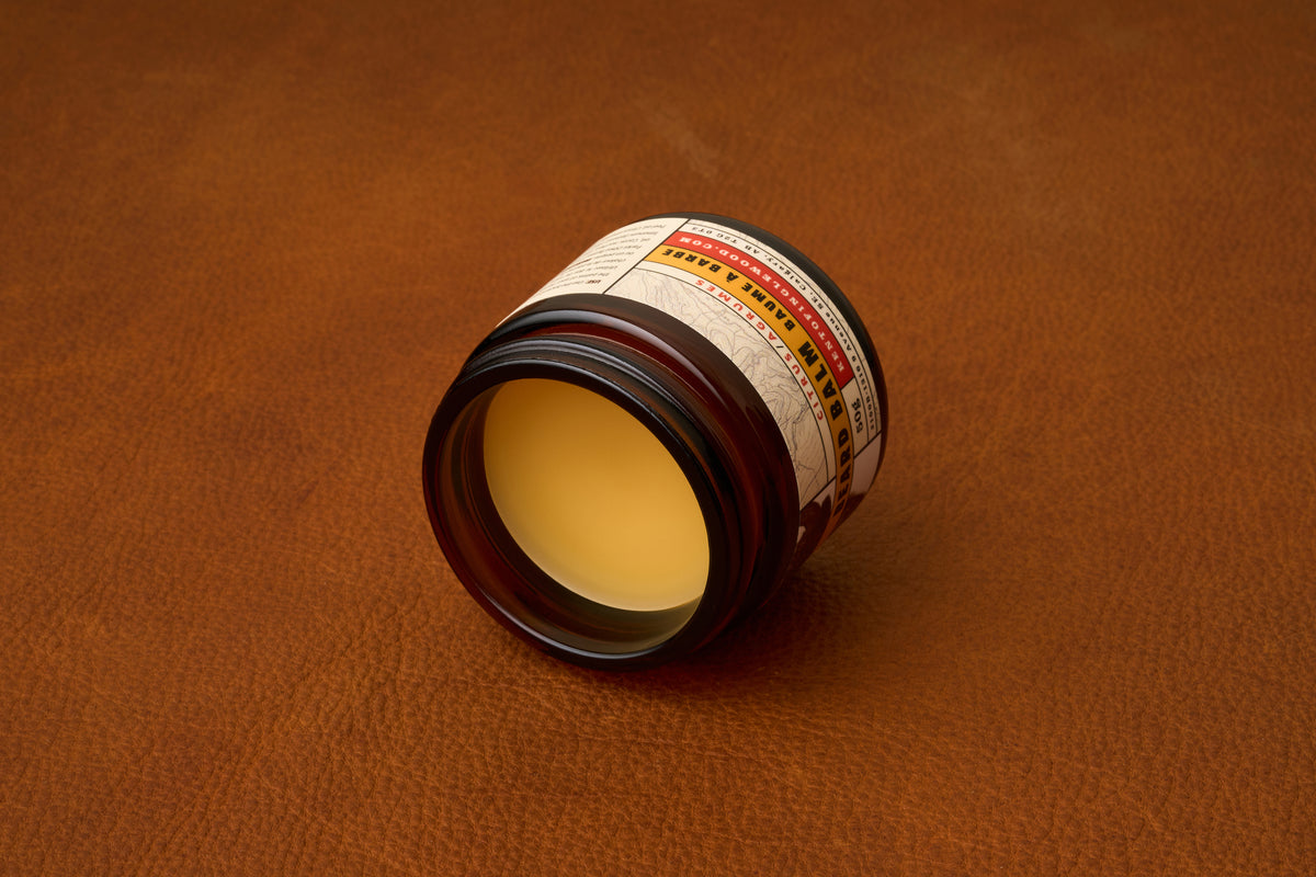 A jar of Kent of Inglewood Citrus Beard Balm lies on its side atop a brown leather surface, its lid removed to reveal the beige balm inside. The brown jar features a white label with text and colored accents.