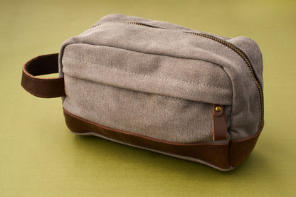 A Kent of Inglewood Dopp Kit, featuring gray canvas and brown leather with a zipper and handle, is displayed on a green surface.