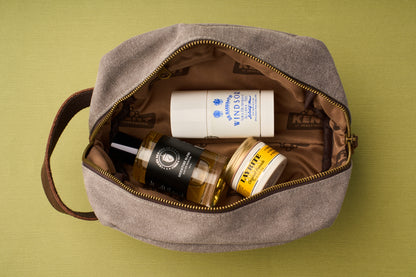 The Kent of Inglewood Dopp Kit—a beige canvas and leather toiletry bag with brown lining—is unzipped to display a bottle of cologne, a white deodorant stick, and a yellow-labeled grooming jar, all set on a green background—making it the ideal choice for stylish dopp kits.
