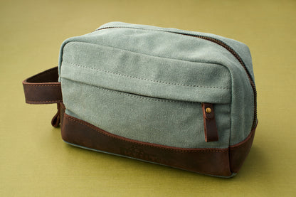 The Kent of Inglewood Dopp Kit, crafted from green canvas with brown leather accents and a handy side handle, sits atop an olive green surface. Ideal for organizing your grooming essentials, this stylish bag features a buttoned front pocket and a visible top zipper—a refined addition to any dopp kit collection.