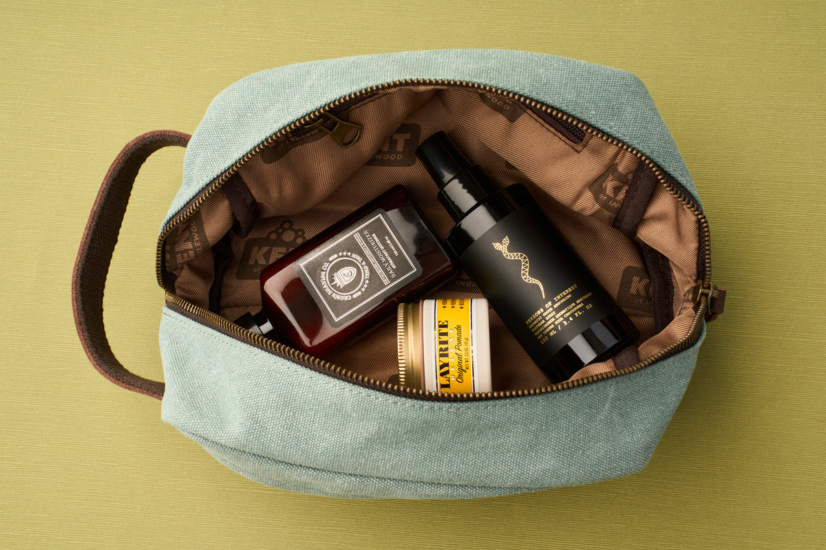 The Kent of Inglewood Dopp Kit, featuring a teal canvas and leather design, is ideal for organizing grooming essentials; inside, a red bottle, black spray bottle, and a yellow-labeled jar are neatly arranged on a green surface.