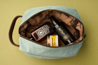 The Kent of Inglewood Dopp Kit, featuring a teal canvas and leather design, is ideal for organizing grooming essentials; inside, a red bottle, black spray bottle, and a yellow-labeled jar are neatly arranged on a green surface.
