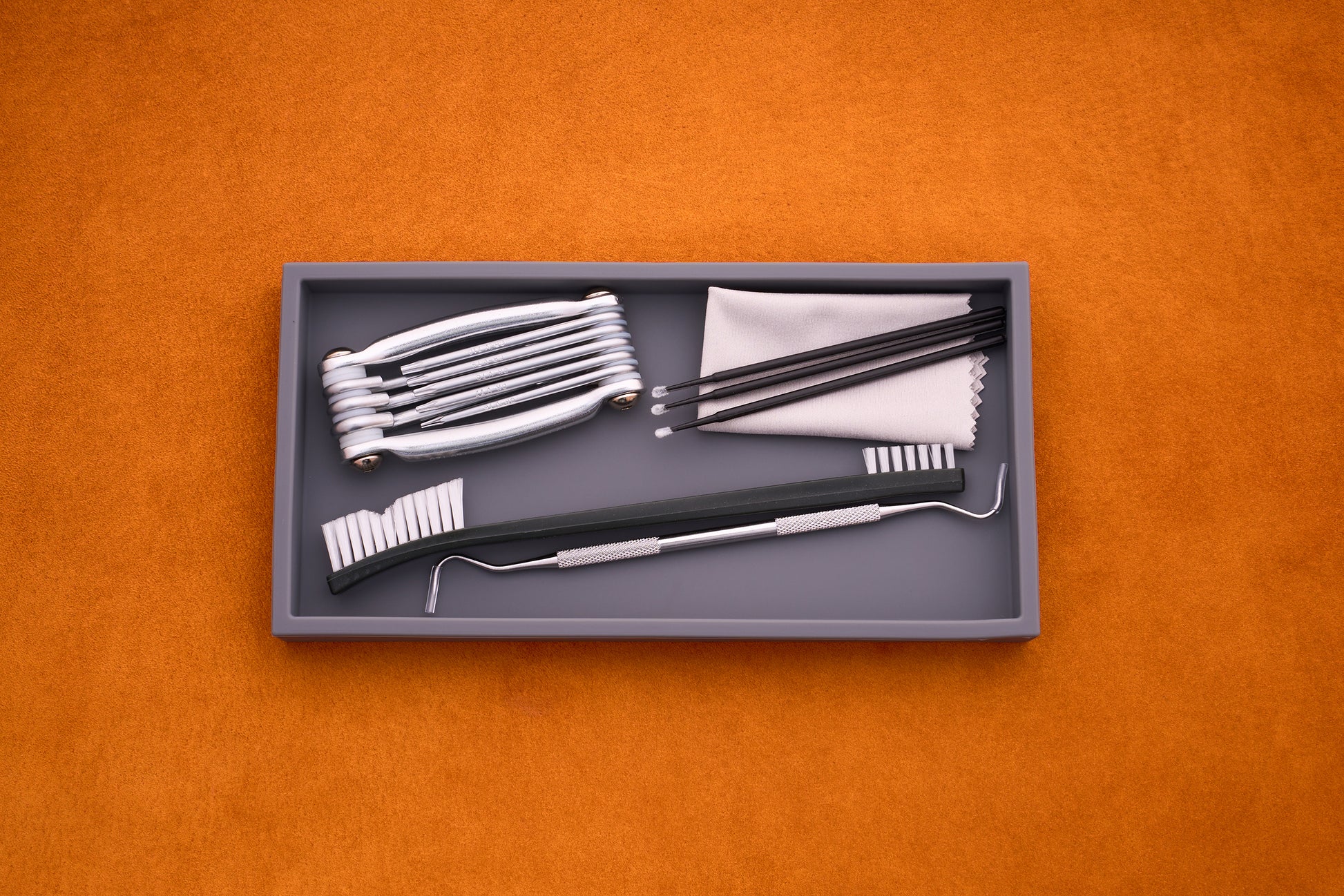 A gray tray on an orange surface holds the Kent of Inglewood Pocket Knife Care Kit, which includes detailing swabs, a brush, scaler, mirrors, tweezers, and a cleaning cloth.