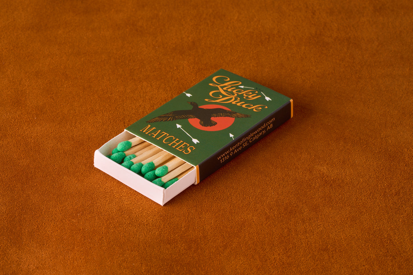Kent of Inglewood Trail Matches in a “Lucky Duck” matchbox, with a flying duck silhouette and green-tipped matches peeking out, on an orange-brown surface.