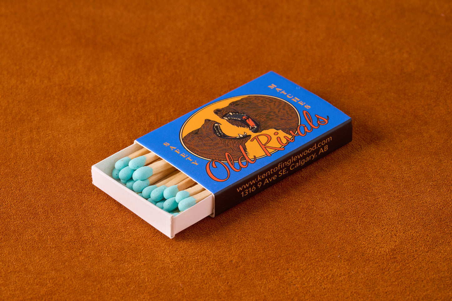 Kent of Inglewood Trail Matches in a bright blue box with snarling bears illustration, showcasing durable wooden matches with turquoise heads on a textured surface.