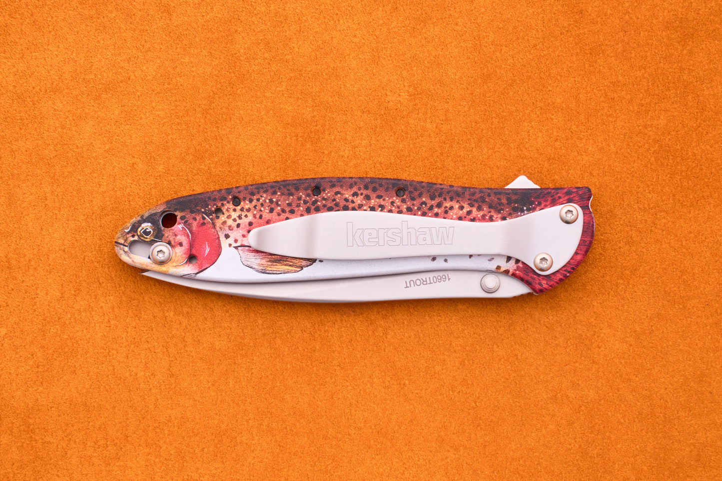The Kershaw Leek Rainbow Trout folding pocket knife, featuring a handle designed to resemble a colorful fish, is displayed on an orange textured surface.