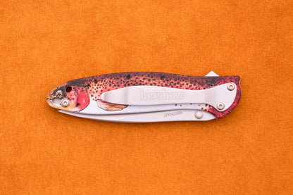 The Kershaw Leek Rainbow Trout folding pocket knife, featuring a handle designed to resemble a colorful fish, is displayed on an orange textured surface.