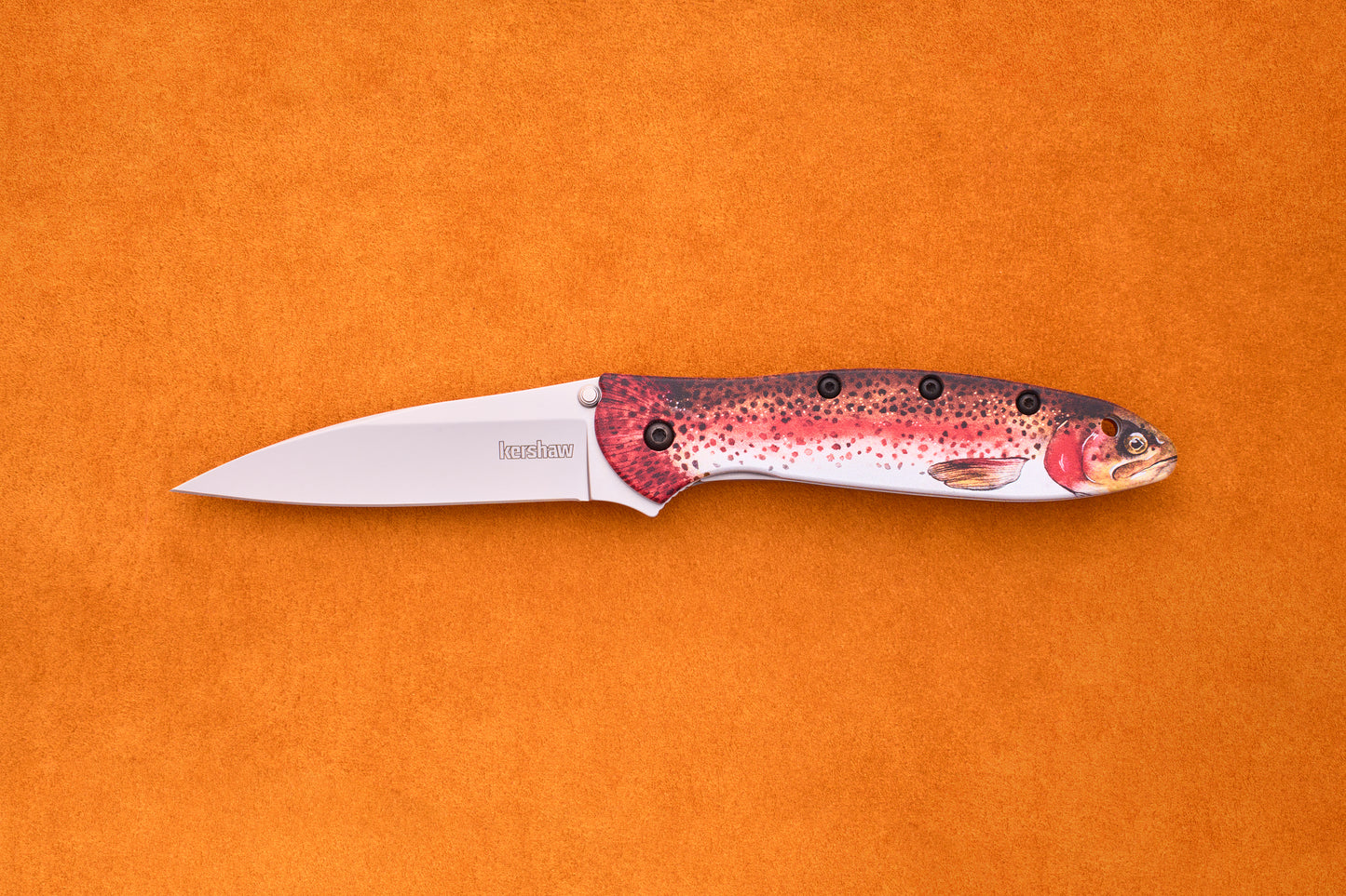 The Kershaw Leek Rainbow Trout, with its silver blade and handle adorned with a vibrant fish motif, is displayed against an orange background—making it a standout EDC knife for any collection.