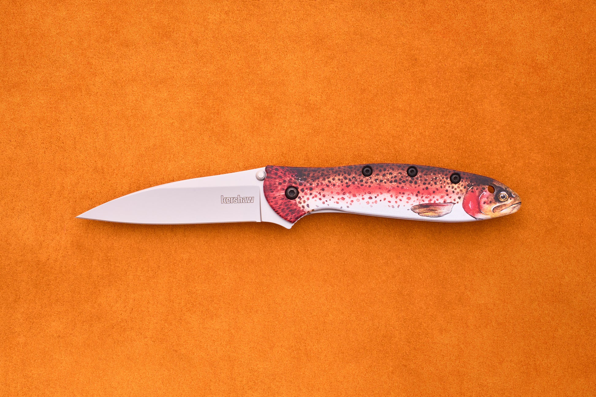The Kershaw Leek Rainbow Trout, with its silver blade and handle adorned with a vibrant fish motif, is displayed against an orange background—making it a standout EDC knife for any collection.