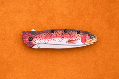 A Kershaw Leek Rainbow Trout folding pocket knife, featuring a handle designed to resemble a colorful fish, shown against an orange background.