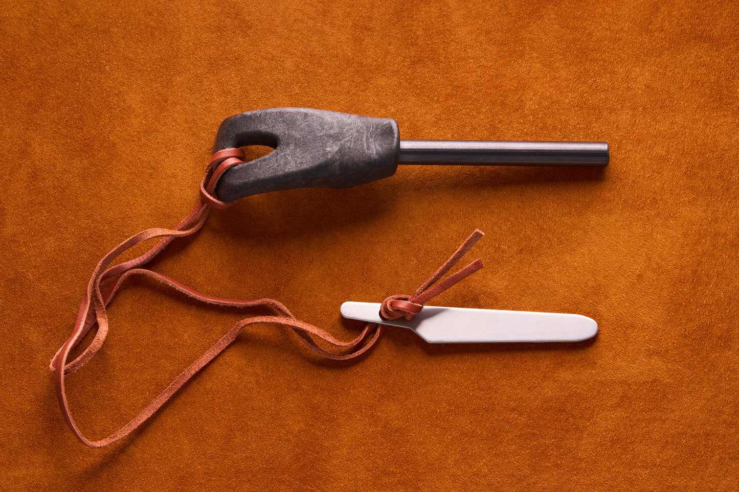 Kupilka Firesteel kit on suede surface, featuring a ferrocerium rod with a molded handle, metal striker blade, and leather thong. Ideal for igniting tinder.