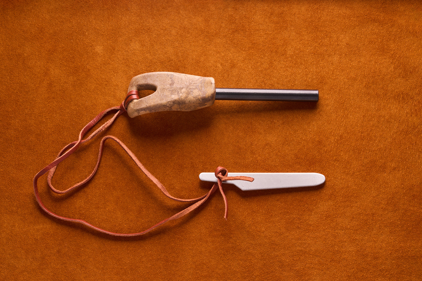 Kupilka Firesteel kit on suede-like surface, featuring a ferro rod with a wooden handle and a metal striker blade, linked by a leather cord.