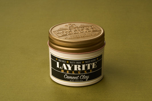 A gold-lidded container of Layrite Cement Hair Clay rests on a plain olive green surface. The label highlights its High Hold, Matte Finish, and notes that it is a water-based pomade ideal for those seeking an extreme hold hair product.