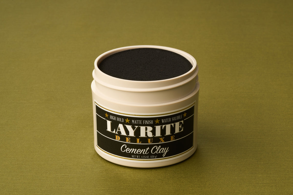 An open white container of Layrite Cement Hair Clay, recognized for its extreme hold, is placed on a green surface. The label is black with gold and white text highlighting the features of this water-based hair clay.