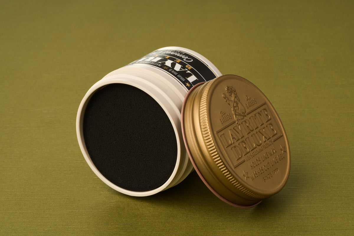 A tin of Layrite Cement Hair Clay with a gold lid, set on its side so the black product inside and the top label are both visible, against an olive green background.