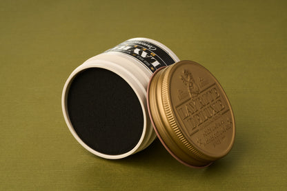 A tin of Layrite Cement Hair Clay with a gold lid, set on its side so the black product inside and the top label are both visible, against an olive green background.