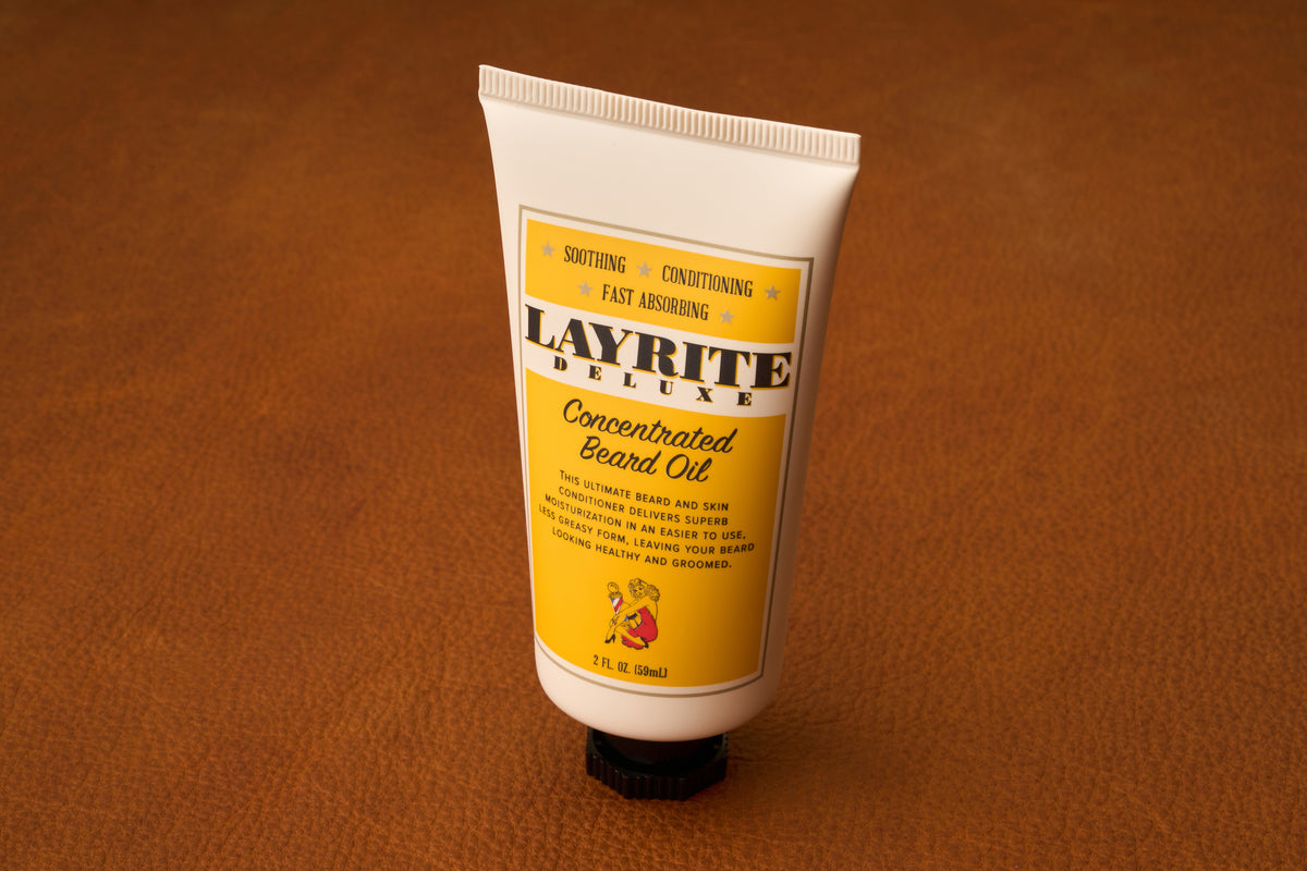 A tube of Layrite Concentrated Beard Oil with a yellow label stands upright on a brown leather surface. The tube, recognized for its effective beard moisturizing properties, displays product details and features a small red lion illustration near the bottom.