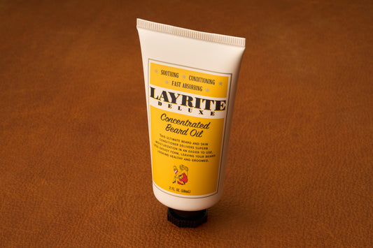 A tube of Layrite Concentrated Beard Oil with a yellow label stands upright on a brown leather surface. The tube, recognized for its effective beard moisturizing properties, displays product details and features a small red lion illustration near the bottom.